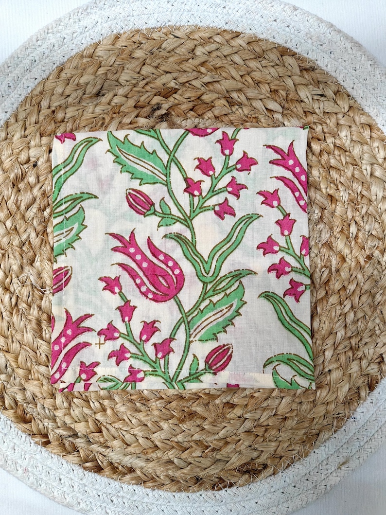May include: Square white fabric with a floral pattern featuring pink flowers and green leaves. The fabric is placed on a round woven jute placemat with a white border. The floral design includes stylized tulips and smaller flower clusters.