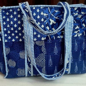 May include: A quilted tote bag in shades of blue and white, featuring various patterns including teardrops, circles, and leaf designs. The bag has sturdy handles and a side pocket, perfect for carrying essentials. The bag is approximately 15 inches wide and 12 inches tall.