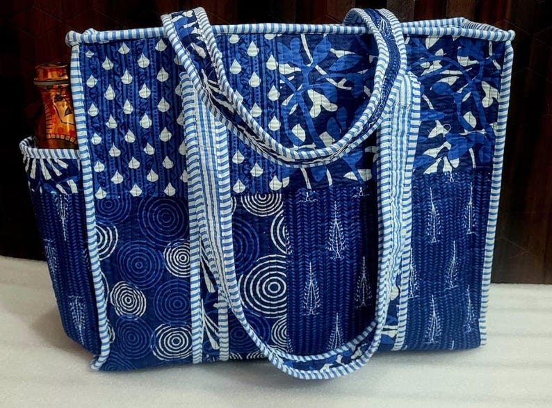 May include: A quilted tote bag in shades of blue and white, featuring various patterns including teardrops, circles, and leaf designs. The bag has sturdy handles and a side pocket, perfect for carrying essentials. The bag is approximately 15 inches wide and 12 inches tall.
