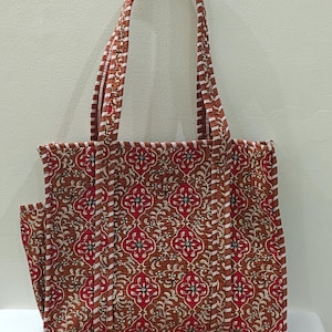 May include: A brown tote bag with a floral pattern in red and white. The bag has a matching handle and a side pocket. The bag is made of quilted fabric and is designed for carrying items.