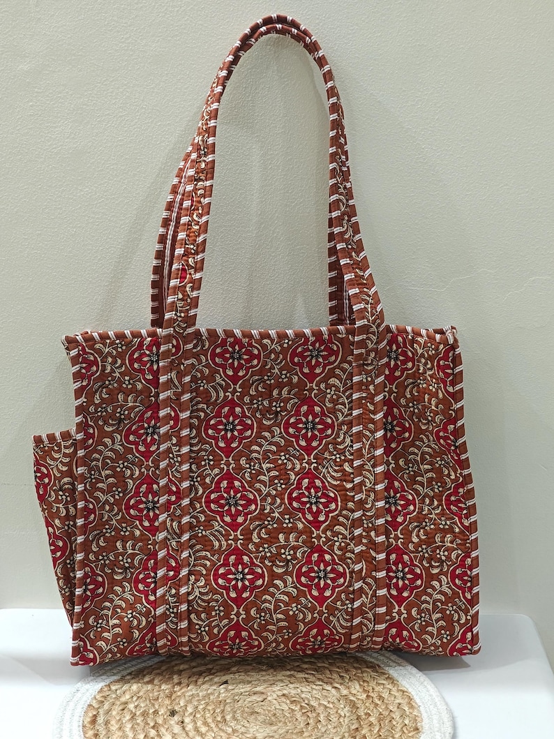 May include: A brown tote bag with a floral pattern in red and white. The bag has a matching handle and a side pocket. The bag is made of quilted fabric and is designed for carrying items.