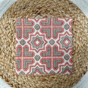May include: A square, patterned textile with a geometric design in shades of pink, gray, and white. The textile is placed on a round, woven jute placemat with a white border. The pattern features interlocking shapes and star-like elements.