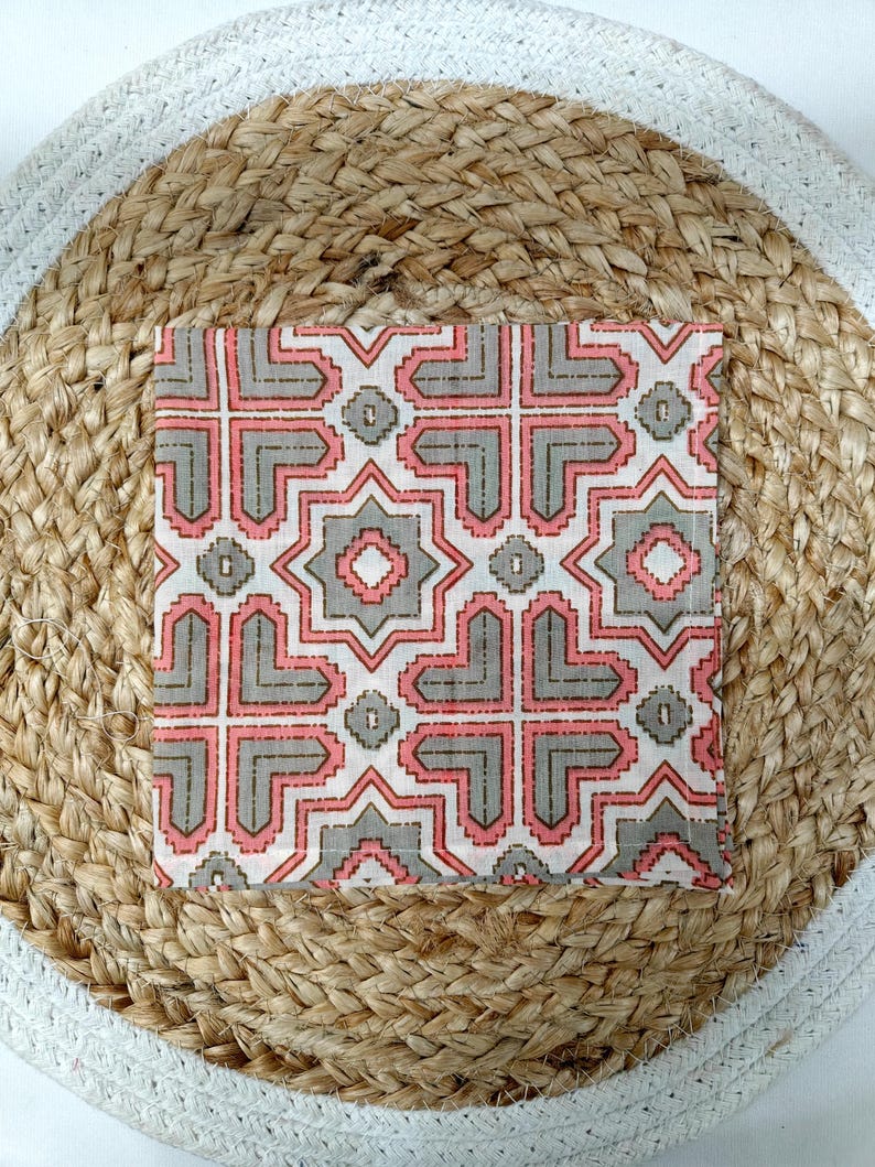 May include: A square, patterned textile with a geometric design in shades of pink, gray, and white. The textile is placed on a round, woven jute placemat with a white border. The pattern features interlocking shapes and star-like elements.