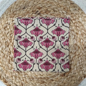 May include: Square beige coaster with a repeating floral pattern in pink and dark gray. The coaster is placed on a round, woven jute placemat with a white border. The floral design features stylized flowers and stems.