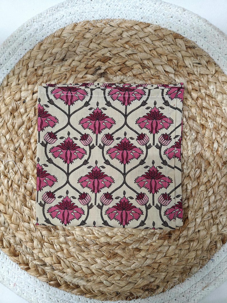 May include: Square beige coaster with a repeating floral pattern in pink and dark gray. The coaster is placed on a round, woven jute placemat with a white border. The floral design features stylized flowers and stems.
