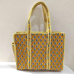 May include: Mustard yellow tote bag with a floral pattern in pink, blue, and white. The bag has a rectangular shape with a side pocket and two handles. The handles and trim are a yellow and white striped fabric.