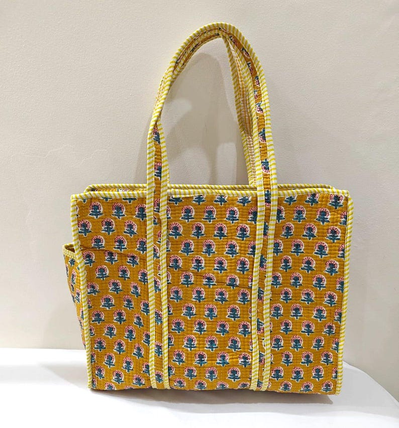 May include: Mustard yellow tote bag with a floral pattern in pink, blue, and white. The bag has a rectangular shape with a side pocket and two handles. The handles and trim are a yellow and white striped fabric.