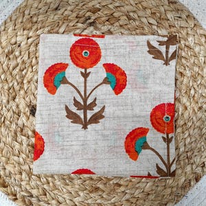 May include: A square, off-white fabric napkin with a repeating floral pattern. The flowers are orange with teal accents and brown stems. The napkin is resting on a woven, round placemat.