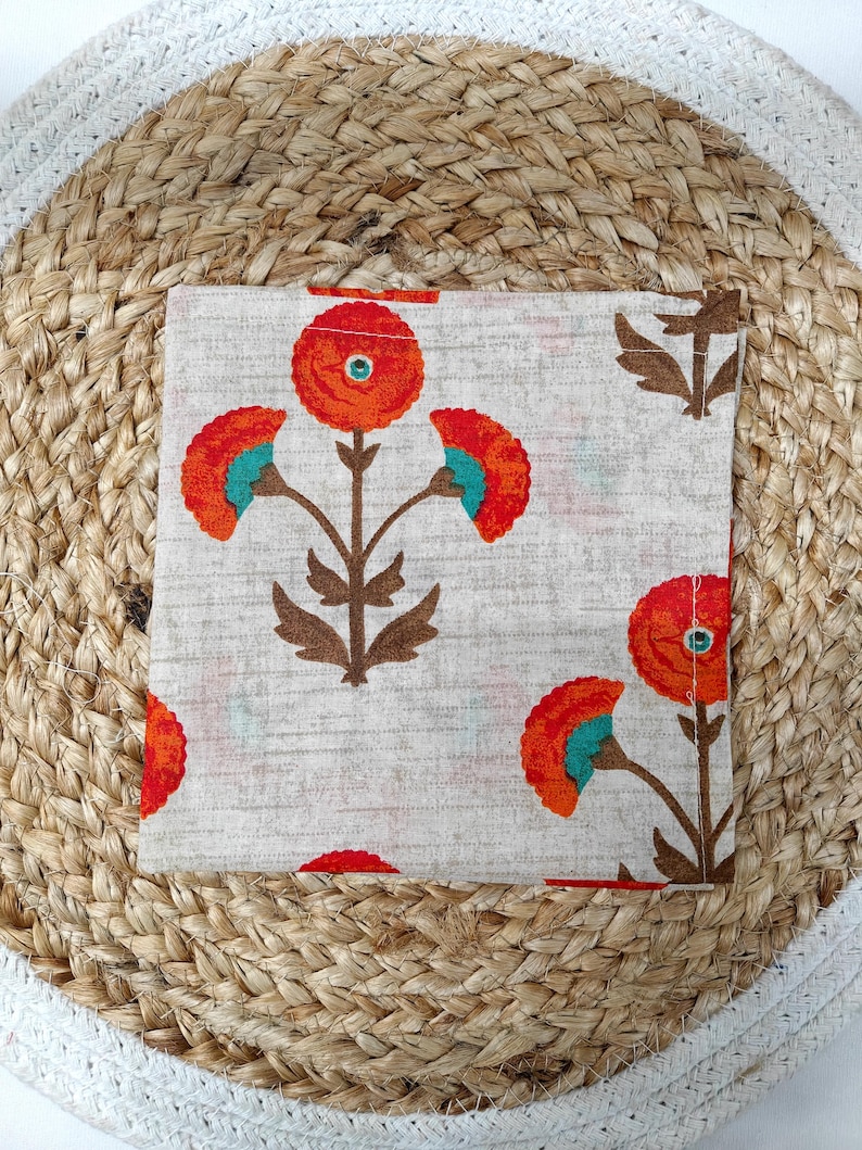 May include: A square, off-white fabric napkin with a repeating floral pattern. The flowers are orange with teal accents and brown stems. The napkin is resting on a woven, round placemat.