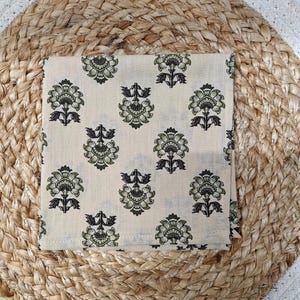 May include: A folded, square, cream-colored cloth with a repeating floral pattern in shades of green and black. The cloth is placed on a woven, circular mat made of natural fibers, creating a textured background.