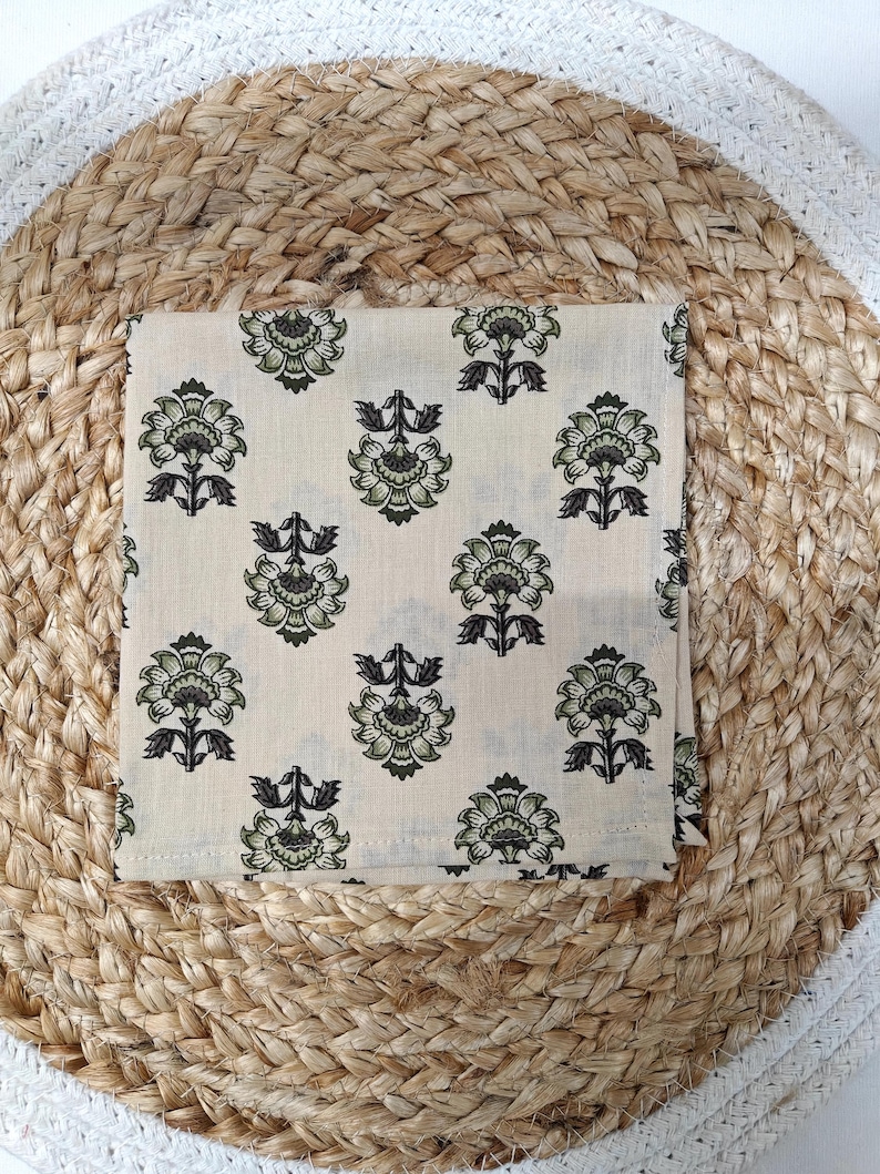 May include: A folded, square, cream-colored cloth with a repeating floral pattern in shades of green and black. The cloth is placed on a woven, circular mat made of natural fibers, creating a textured background.