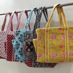 May include: A row of five quilted tote bags hanging on a metal rod. Each bag features a distinct block-printed floral or geometric pattern in vibrant colours including yellow, black, light blue, red, and white, with matching fabric handles.