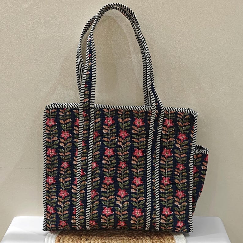 May include: A navy blue tote bag with a floral pattern in pink and green. The bag has a striped trim and handles. The bag is made of quilted fabric and has a rectangular shape.