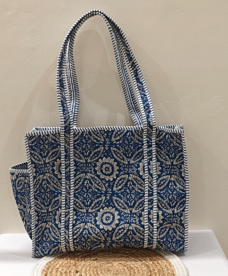 May include: A blue tote bag with a floral pattern and white trim. The bag has a rectangular shape with a side pocket and two handles. The bag is made of a quilted fabric.