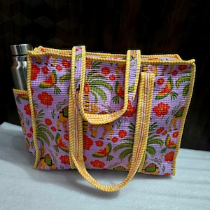 May include: A quilted tote bag with a tropical pattern featuring parrots, palm trees, and flowers in shades of purple, yellow, and red. The bag has a side pocket holding a stainless steel water bottle and yellow handles.