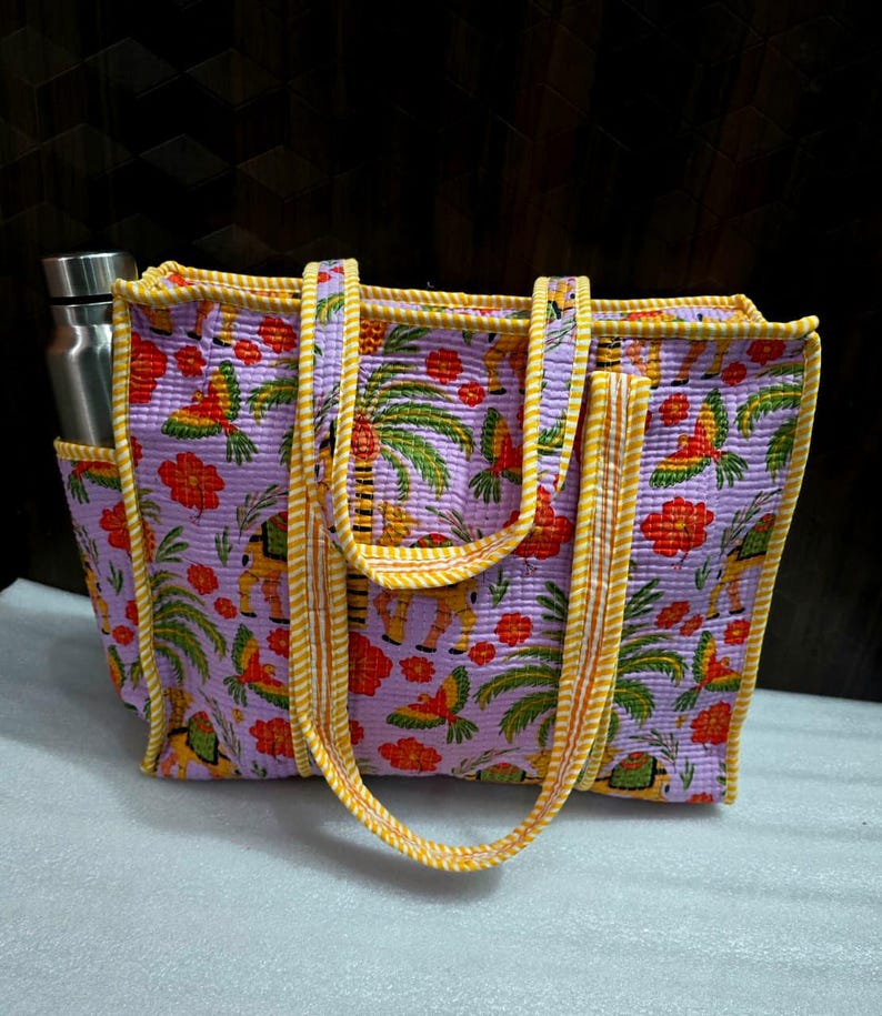May include: A quilted tote bag with a tropical pattern featuring parrots, palm trees, and flowers in shades of purple, yellow, and red. The bag has a side pocket holding a stainless steel water bottle and yellow handles.