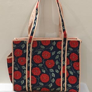 May include: A floral tote bag with a navy blue background and red flower pattern. The bag has a rectangular shape with a side pocket and long handles. The handles and edges are trimmed with a striped fabric.
