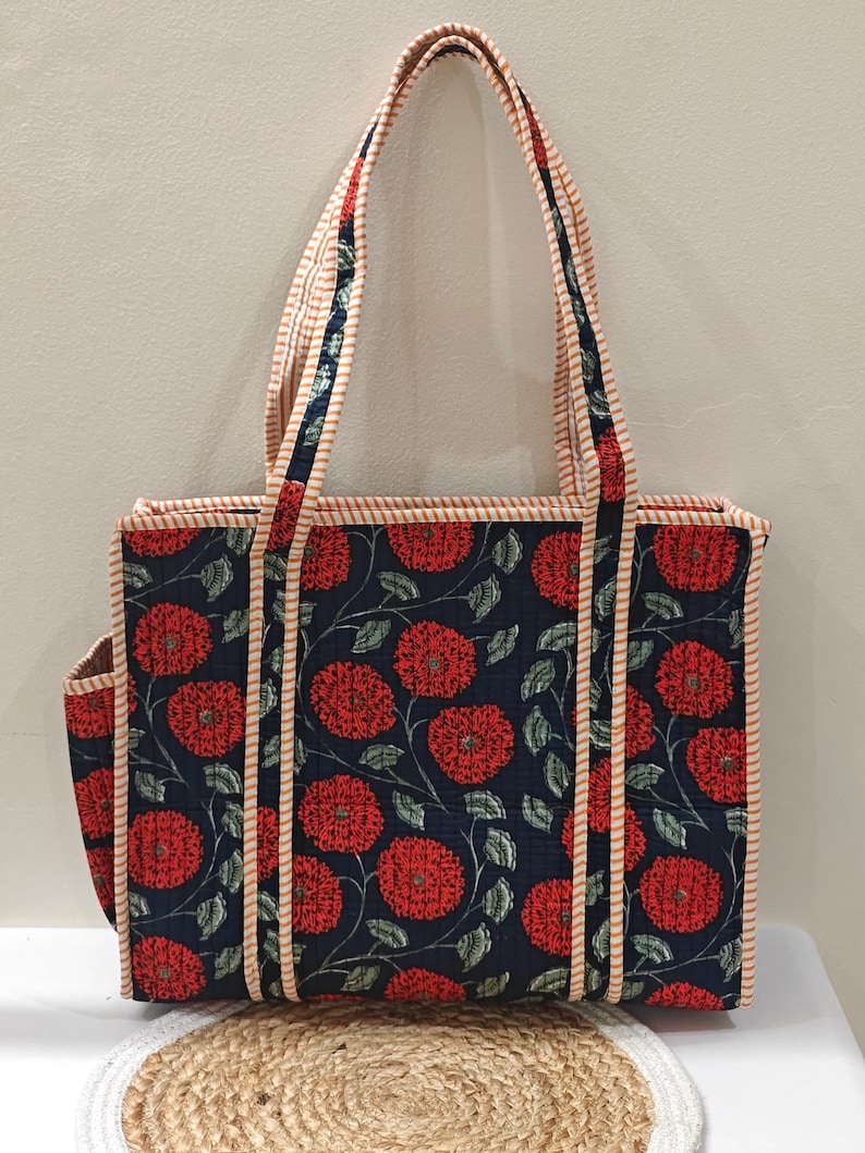 May include: A floral tote bag with a navy blue background and red flower pattern. The bag has a rectangular shape with a side pocket and long handles. The handles and edges are trimmed with a striped fabric.