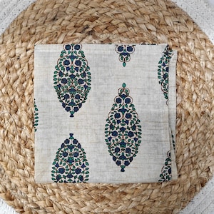 May include: A folded, off-white cloth napkin with a repeating pattern of blue and green floral designs. The napkin is placed on a woven, round placemat made of natural fibers. The placemat has a light brown and white color scheme.
