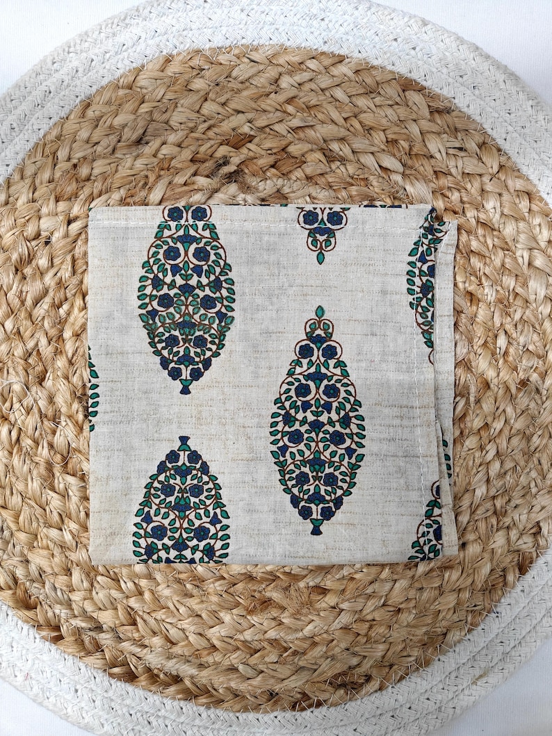 May include: A folded, off-white cloth napkin with a repeating pattern of blue and green floral designs. The napkin is placed on a woven, round placemat made of natural fibers. The placemat has a light brown and white color scheme.