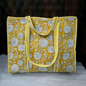 May include: A yellow tote bag with a white floral pattern. The bag has two handles and is made of quilted fabric. The edges and handles are trimmed with a yellow and white striped fabric.