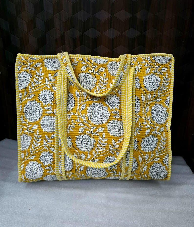May include: A yellow tote bag with a white floral pattern. The bag has two handles and is made of quilted fabric. The edges and handles are trimmed with a yellow and white striped fabric.