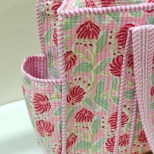 May include: Pink quilted tote bag with a floral pattern in red and green. The bag has a striped trim and handles. The side pocket is also trimmed with stripes. The bag is made of a soft, quilted fabric.