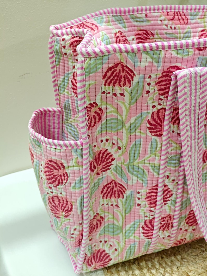 May include: Pink quilted tote bag with a floral pattern in red and green. The bag has a striped trim and handles. The side pocket is also trimmed with stripes. The bag is made of a soft, quilted fabric.