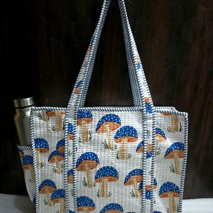 May include: A white quilted tote bag with a repeating pattern of blue and orange mushrooms. The bag has a side pocket and a matching handle with blue and white striped trim. A stainless steel water bottle is in the side pocket.