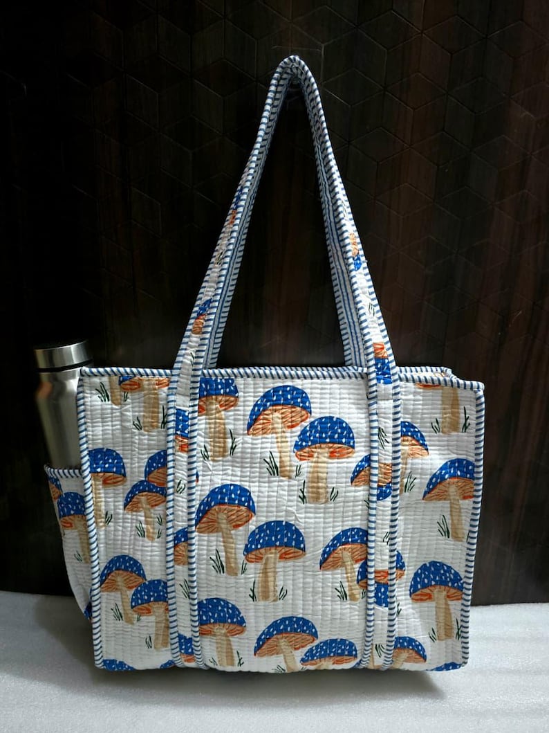 May include: A white quilted tote bag with a repeating pattern of blue and orange mushrooms. The bag has a side pocket and a matching handle with blue and white striped trim. A stainless steel water bottle is in the side pocket.