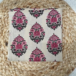 May include: A folded cream-colored fabric with a repeating floral pattern in shades of pink and black. The floral design is symmetrical and stylized. The fabric is resting on a woven, circular mat.