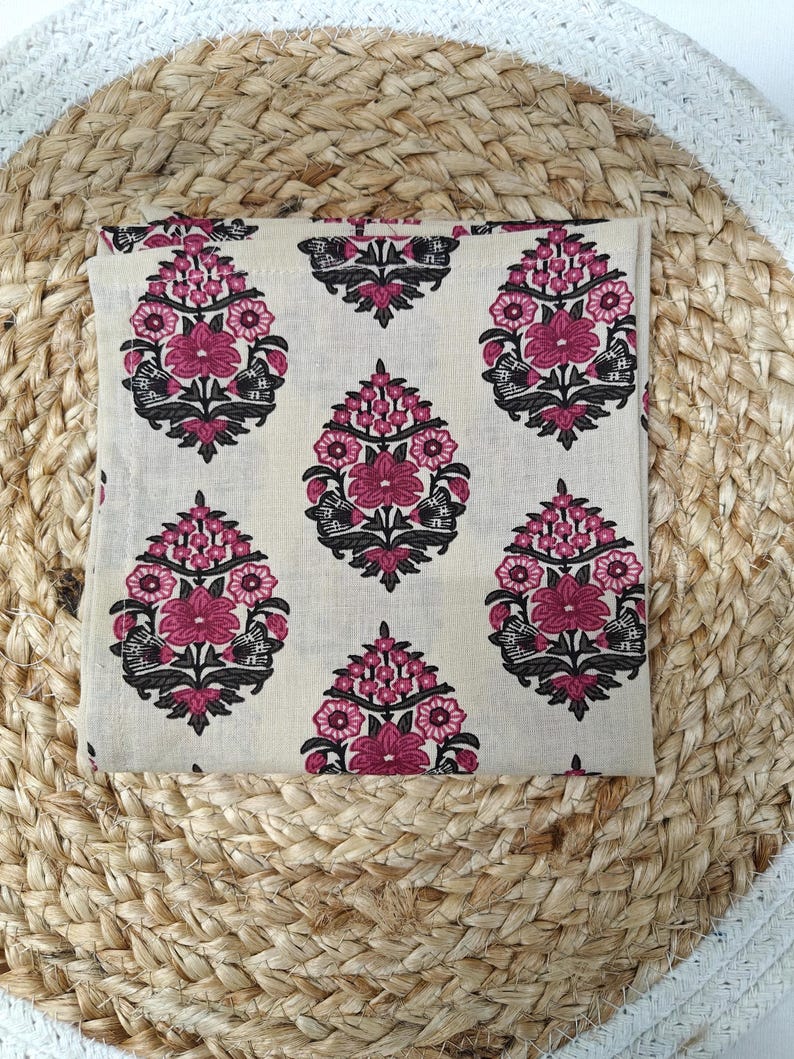 May include: A folded cream-colored fabric with a repeating floral pattern in shades of pink and black. The floral design is symmetrical and stylized. The fabric is resting on a woven, circular mat.