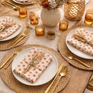 May include: A beautifully set table with woven placemats, white plates, and gold cutlery. Floral patterned napkins are tied with twine. A vase of dried flowers and candles complete the warm, inviting dining scene.