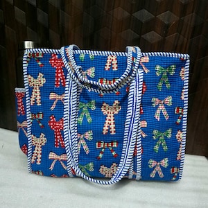 May include: A blue tote bag with a repeating pattern of red, green, and white bows. The bag has a white and blue striped trim and handles. A water bottle is visible in the side pocket.