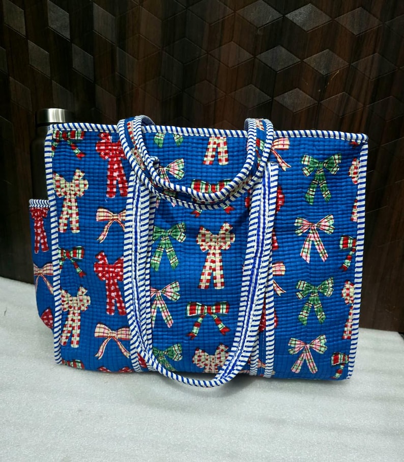May include: A blue tote bag with a repeating pattern of red, green, and white bows. The bag has a white and blue striped trim and handles. A water bottle is visible in the side pocket.