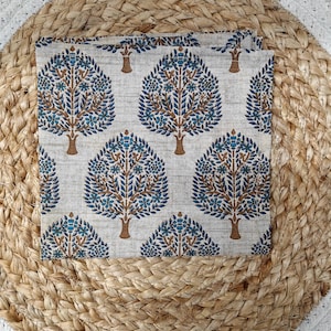 May include: A square, folded fabric with a repeating tree pattern. The trees have brown trunks and branches with blue and teal leaves and flowers. The fabric is set on a woven, beige placemat.