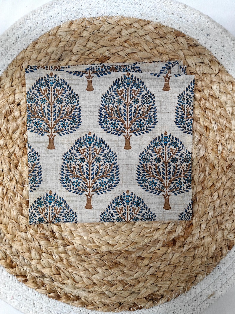 May include: A square, folded fabric with a repeating tree pattern. The trees have brown trunks and branches with blue and teal leaves and flowers. The fabric is set on a woven, beige placemat.