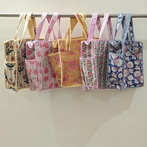 May include: A selection of patterned tote bags hanging on a rail. The bags feature floral designs in various colours, including pink, yellow, and blue. Each bag has a unique pattern, with matching handles and external pockets.