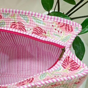 May include: A quilted tote bag with a pink floral pattern and a striped interior. The bag features a bright pink zipper and a white and pink striped trim. The bag is made of a soft, quilted fabric.
