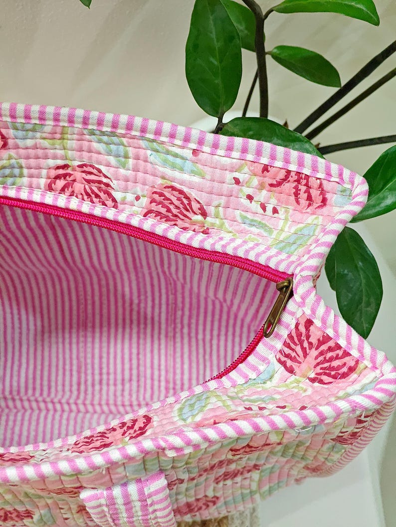 May include: A quilted tote bag with a pink floral pattern and a striped interior. The bag features a bright pink zipper and a white and pink striped trim. The bag is made of a soft, quilted fabric.