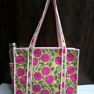 May include: A floral tote bag with a pink and green pattern. The bag has a light yellow background with large pink flowers and green vines. The edges and handles are trimmed in pink. A stainless steel water bottle is in the side pocket.