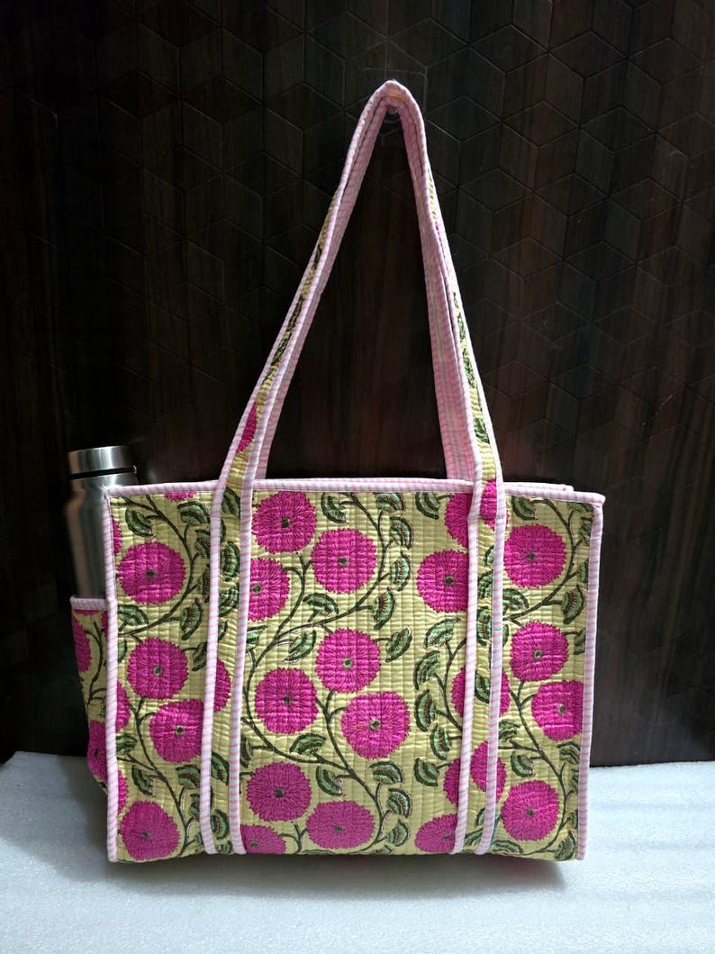 May include: A floral tote bag with a pink and green pattern. The bag has a light yellow background with large pink flowers and green vines. The edges and handles are trimmed in pink. A stainless steel water bottle is in the side pocket.