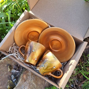 May include: A set of two ceramic coffee cups and saucers in a cardboard box. The cups and saucers are a warm, golden color with a glossy finish. Each cup has a decorative design of white hearts and lines. The box is filled with shredded paper.