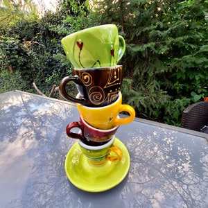 May include: A stack of four colorful ceramic coffee cups with unique designs. The cups are stacked on a green saucer and are on a gray table with a reflection of trees.