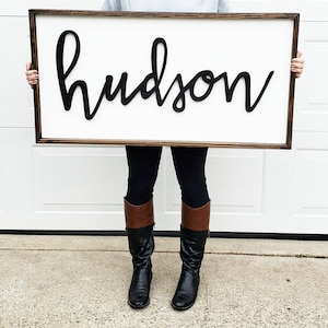 May include: A white wooden sign with a brown frame, featuring the name "hudson" in black, 3D script lettering.
