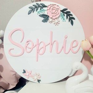 Peut inclure: Une pancarte ronde en bois blanc avec le nom "Sophie" écrit en lettres cursives roses. La pancarte est décorée de fleurs et de feuilles en papier roses et vertes.