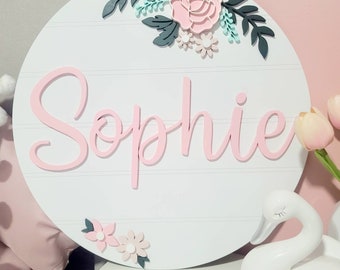 以下が含まれることがあります： ピンクの筆記体で「Sophie」と書かれた白い丸い木製の看板。看板はピンクと緑の紙の花と葉で飾られています。