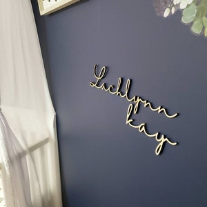 May include: Wooden cursive name sign spelling "Lachlynn Kay" in a gold color, mounted on a dark blue wall. The sign is a decorative element, possibly for a nursery or bedroom. A white curtain is visible on the left.