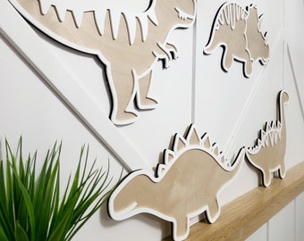 May include: Four wooden dinosaur wall decorations in various poses. The dinosaurs are painted in a light brown color with white outlines. The dinosaurs are mounted on a white wall with a wood-grain pattern.