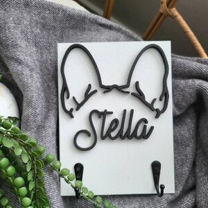 May include: White wooden key holder with a black silhouette of a French bulldog's head and the name "Stella" in black lettering. Two black hooks are attached to the bottom of the holder.
